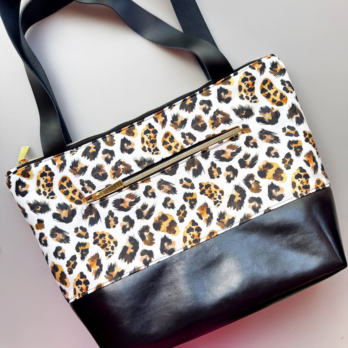 A leopard print tote bag with black faux-leather trim and handles, featuring a gold-tone zipper on the front pocket, photographed on a light gray background.