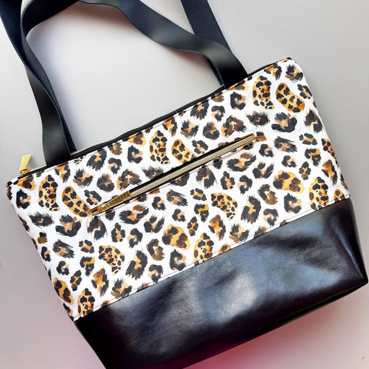 A leopard print tote bag with black faux-leather trim and handles, featuring a gold-tone zipper on the front pocket, photographed on a light gray background.