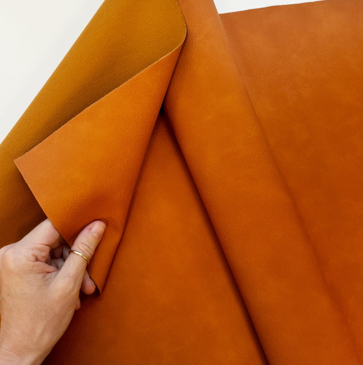 A close-up of a hand holding a folded piece of warm orange-brown leather or faux leather fabric, showing its smooth texture and color against a plain light background.
