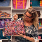 A woman wearing glasses and a patterned top holds up a colorful floral fabric tote bag with a black and white striped handle in a sewing and bagging studio. Shelves behind her are filled with various colorful bags and sewing supplies.