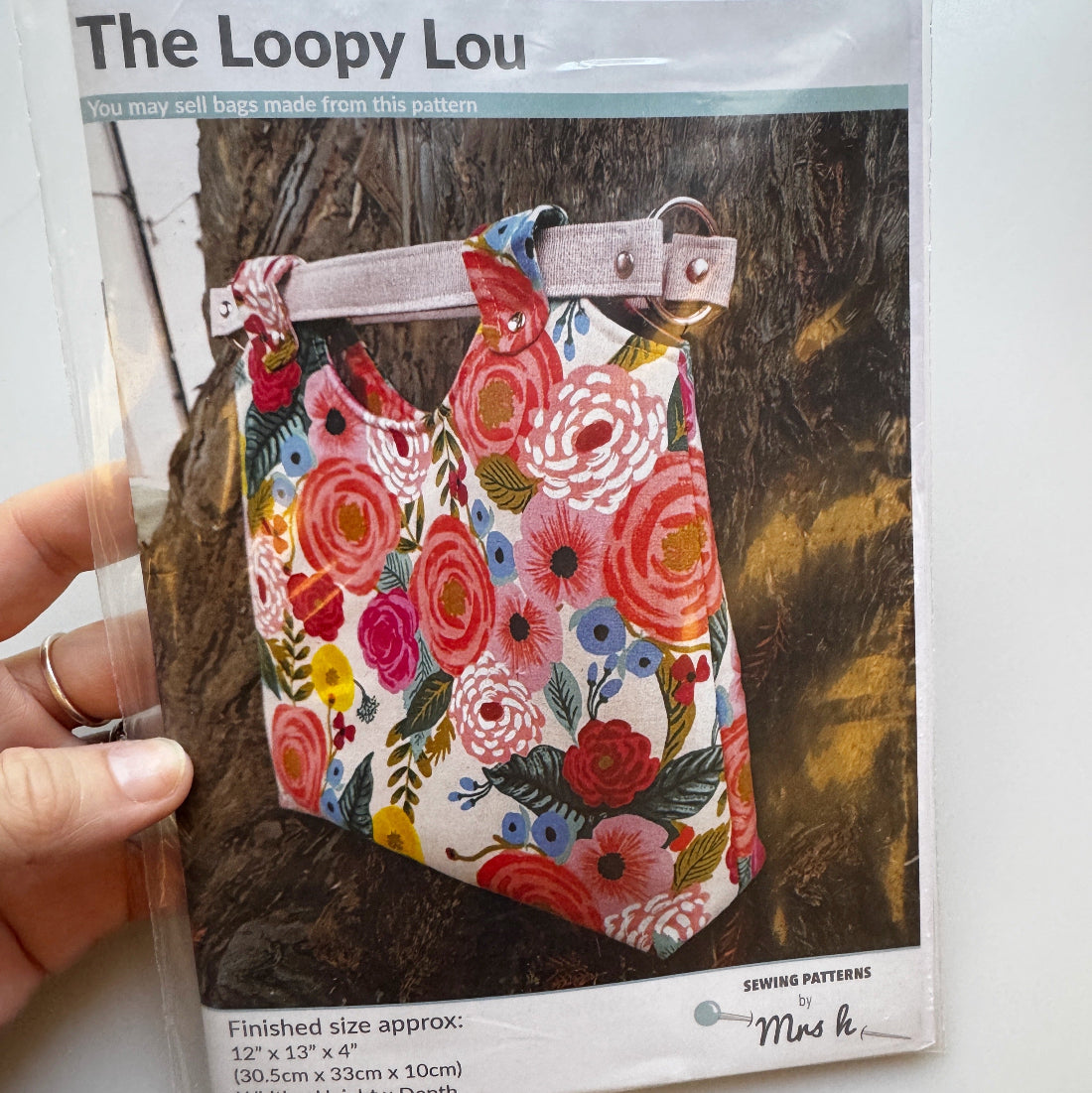 A hand holds a clear plastic bag containing a sewing pattern for 'The Loopy Lou' pet bag. The pattern cover shows a colorful floral print dog bag with a light brown strap and metal ring, set against a dark wood background.