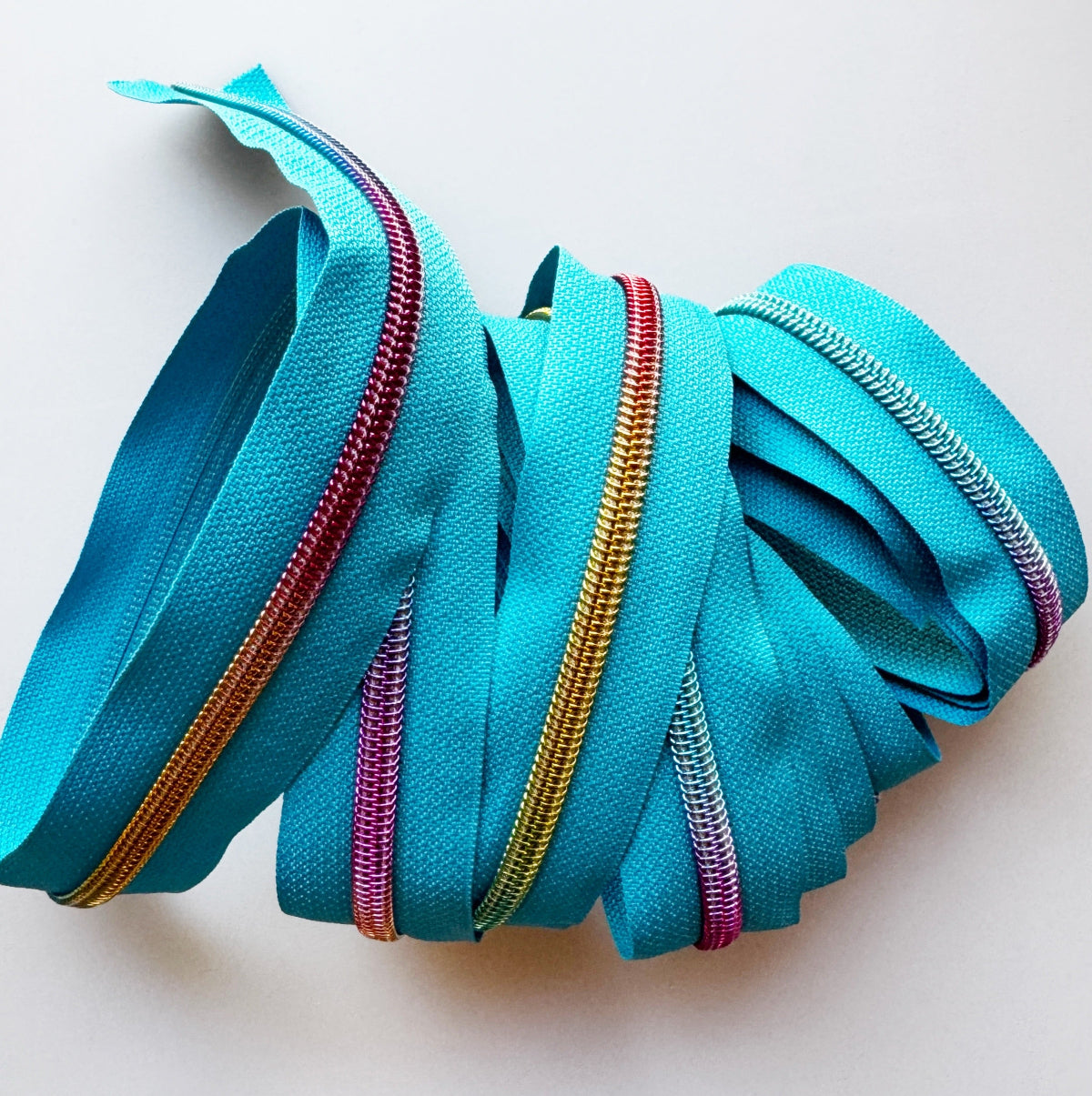 A coiled bundle of turquoise blue zipper tape or ribbon with metallic zipper edges in red, gold, and silver, arranged on a plain light background.