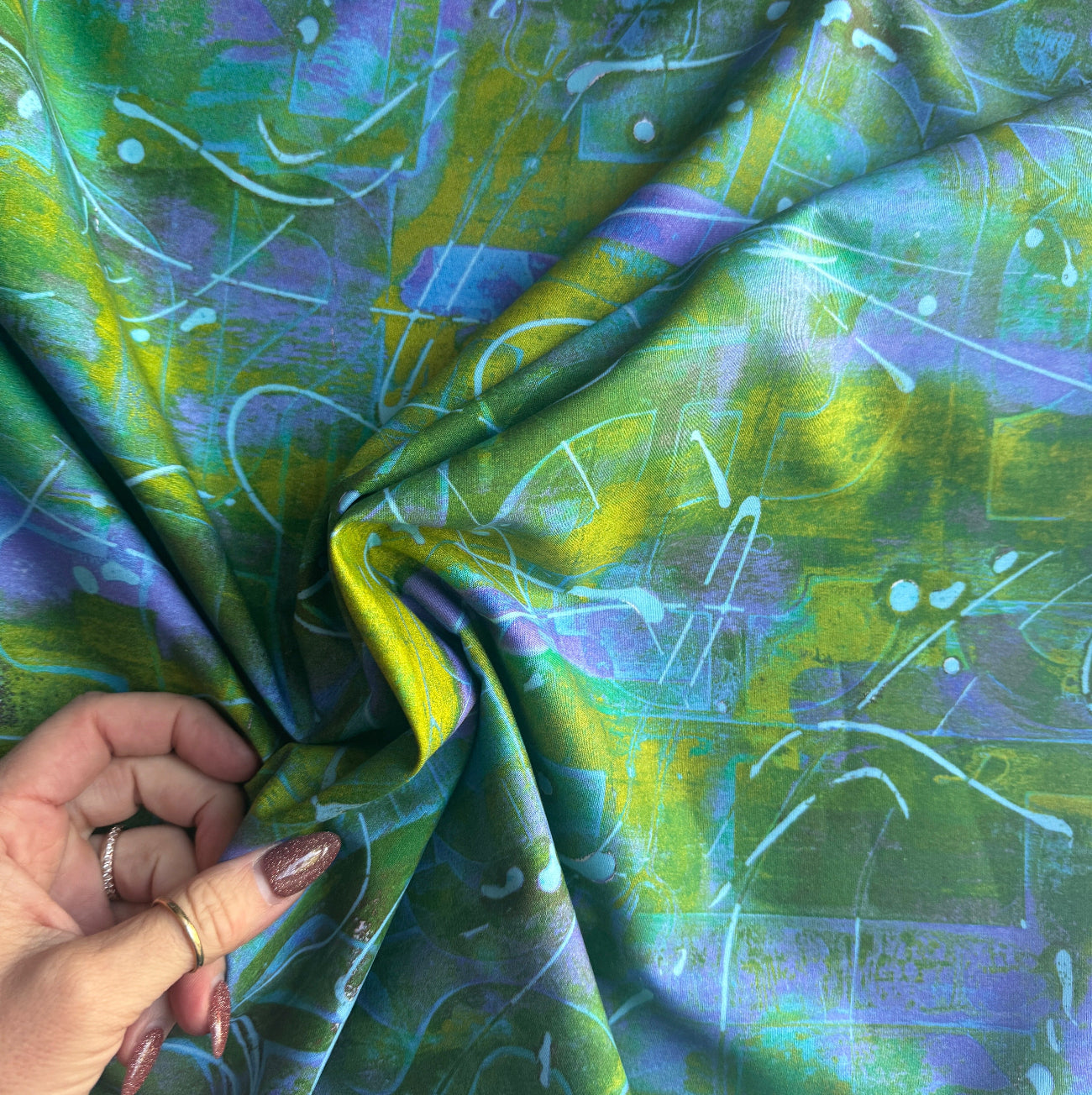 A hand gathering a section of vibrant green, yellow, and purple abstract print fabric to show its soft drape and texture.