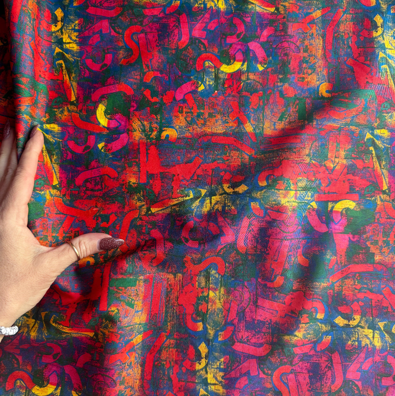 A hand holding a section of colorful, abstract-print fabric, showing the material's drape, fine weave texture, and vibrant red and blue palette.
