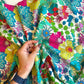 A close-up shot of a hand gently holding a section of Sally Kelly botanical floral cotton fabric sold by the yard, providing a helpful sense of pattern scale for quilting and sewing projects. The intricate textile print displays whimsical, multi-colored flowers and wildflower pods in rich tones of hot pink, yellow, teal, and kelly green. The hand wears a dark blue watch and gold rings against the bright, draped fabric.