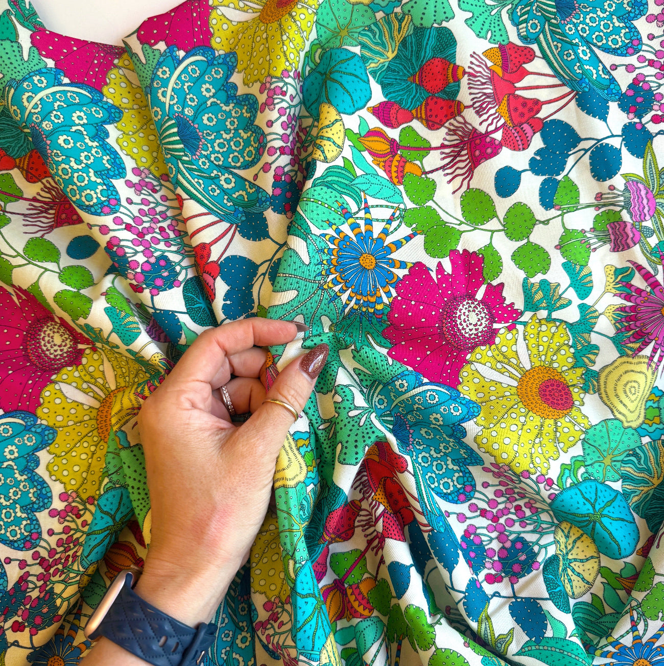 A close-up shot of a hand gently holding a section of Sally Kelly botanical floral cotton fabric sold by the yard, providing a helpful sense of pattern scale for quilting and sewing projects. The intricate textile print displays whimsical, multi-colored flowers and wildflower pods in rich tones of hot pink, yellow, teal, and kelly green. The hand wears a dark blue watch and gold rings against the bright, draped fabric.