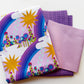 Three folded cotton quilting fabrics from the Alison Glass Postmark collection arranged on a white background. The top fabric features a whimsical light lavender print with bright yellow suns, purple and indigo rainbows, white clouds, and colorful teal, magenta, and green flowers with raindrops. Coordinating deep purple crosshatch grid fabric and soft mauve fabric with tiny dark purple dots complete this colorful fabric bundle for quilting, pillowcases, and handmade sewing projects.