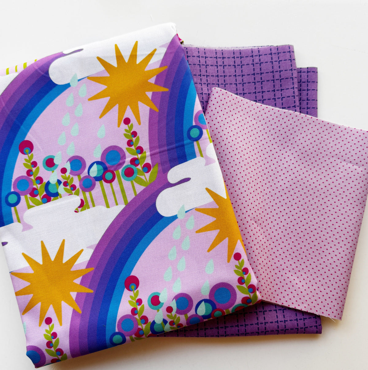 Three folded cotton quilting fabrics from the Alison Glass Postmark collection arranged on a white background. The top fabric features a whimsical light lavender print with bright yellow suns, purple and indigo rainbows, white clouds, and colorful teal, magenta, and green flowers with raindrops. Coordinating deep purple crosshatch grid fabric and soft mauve fabric with tiny dark purple dots complete this colorful fabric bundle for quilting, pillowcases, and handmade sewing projects.