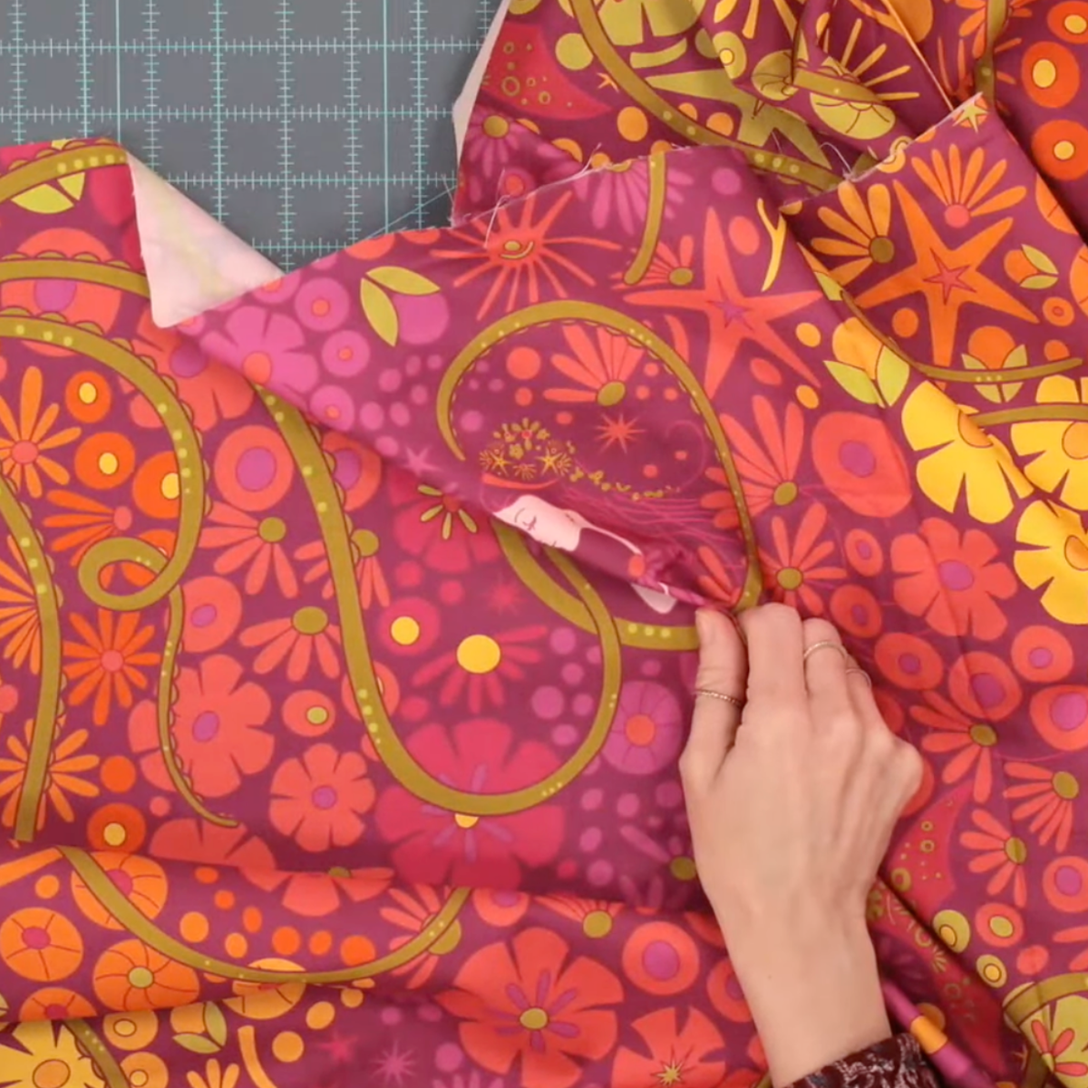 Alison Glass Redwood 108-inch Bloom cotton quilting fabric, SKU 10462-E, featuring a vibrant retro psychedelic print with magenta, hot pink, orange, and golden yellow flowers, sunbursts, and paisley shapes, displayed on a green cutting mat