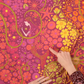 Close-up of Alison Glass Redwood 108-inch Bloom cotton quilting fabric featuring psychedelic pink, magenta, orange, and golden yellow flowers, swirling paisley shapes, and a woman holding a glowing star, with hands highlighting the colorful floral print.