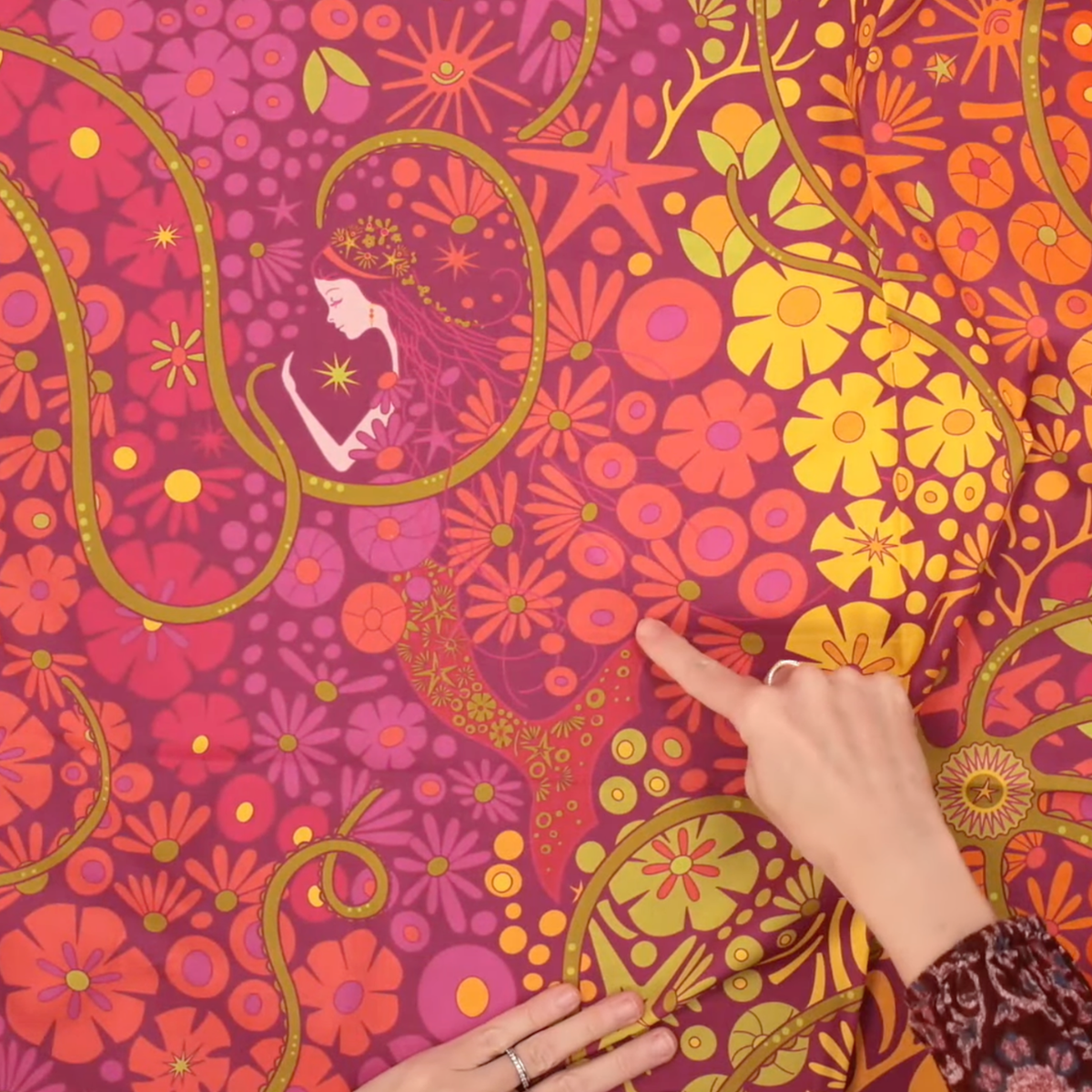 Close-up of Alison Glass Redwood 108-inch Bloom cotton quilting fabric featuring psychedelic pink, magenta, orange, and golden yellow flowers, swirling paisley shapes, and a woman holding a glowing star, with hands highlighting the colorful floral print.