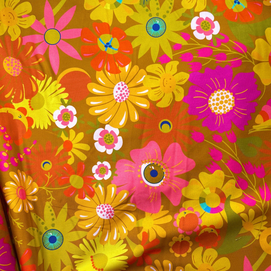 A close-up of a vibrant, multicolored floral fabric with a retro 1970s pattern. A hand is visible at the bottom left, holding the fabric to show its drape and texture.
