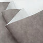 A close-up of a rolled piece of textured faux leather or vinyl material in a warm gray-brown tone, showing both the outer textured surface and the smoother backing against a plain white background.