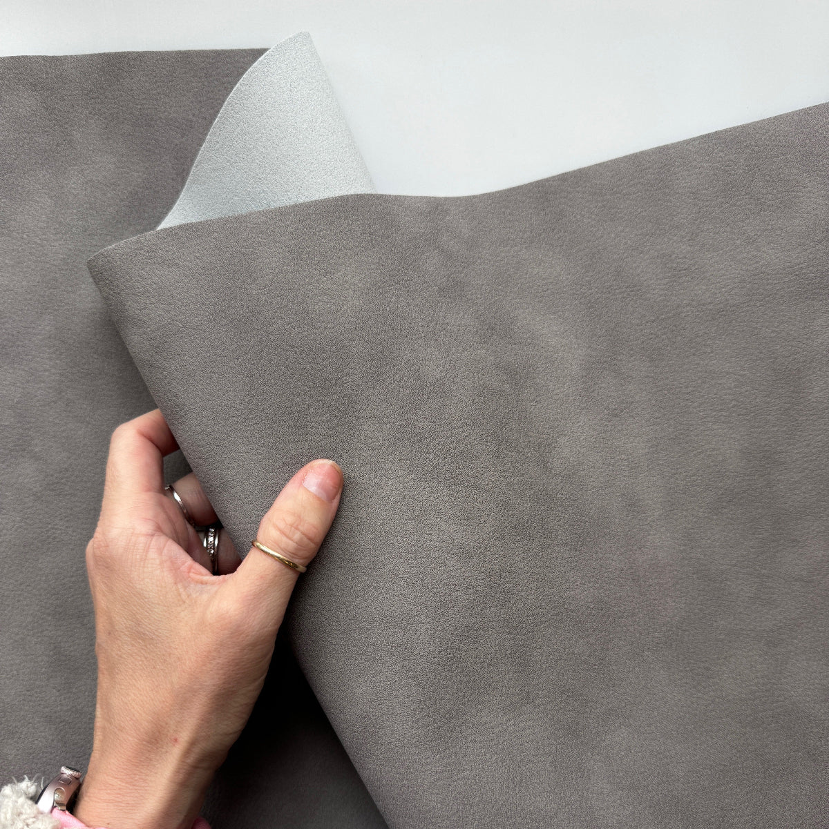 A close-up of a person's hand lifting a corner of a large gray textured material, likely a sheet of vinyl, contact paper, or leather-like fabric, showing its matte finish and reverse side against a plain light background.