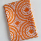 A neatly folded piece of Treasure Splendour Tang cotton fabric from the Indigo and Aster collection by Bari J for Art Gallery Fabrics, showing a bright orange floral medallion print on a soft light pink background.