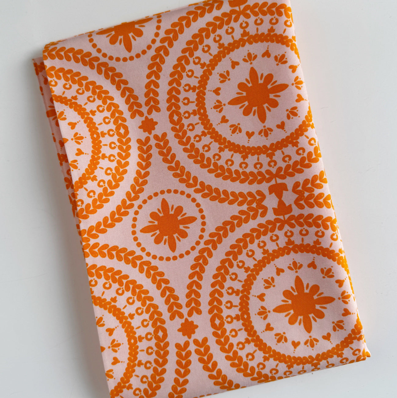 A neatly folded piece of Treasure Splendour Tang cotton fabric from the Indigo and Aster collection by Bari J for Art Gallery Fabrics, showing a bright orange floral medallion print on a soft light pink background.