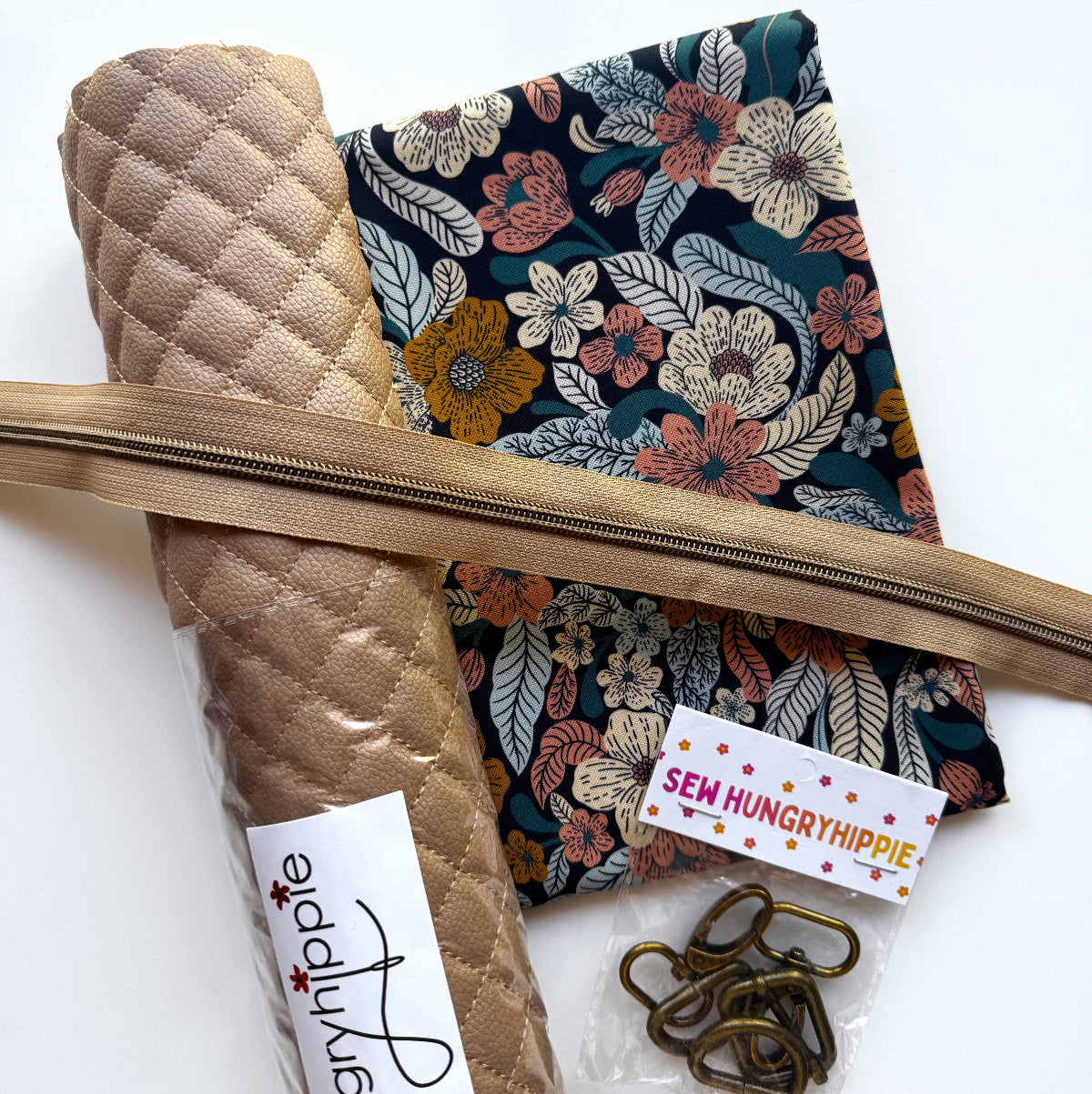 A sewing kit laid out on a white background, including a rolled tan quilted fabric piece, a dark floral fabric panel, a tan zipper, metal clips, and a small branded tag reading 'Sem Hungry Hippie'.