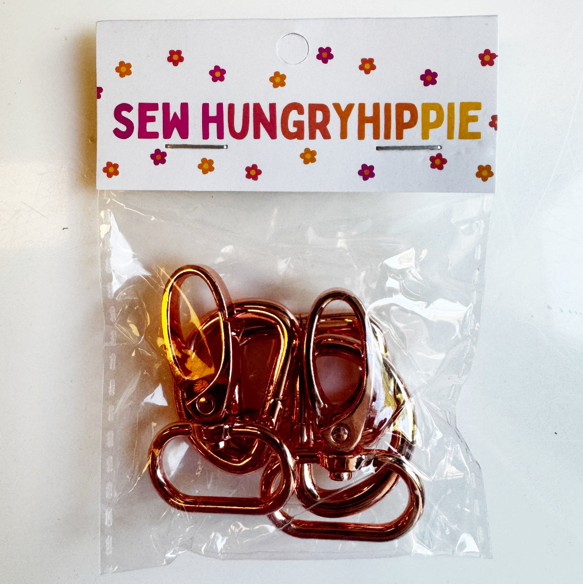 A clear plastic bag containing several metallic rose gold sewing clips, sealed with a white paper header card printed with the brand name 'SEW HUNGRYHIPPIE' in colorful letters and small flower illustrations.