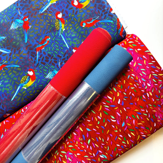 Three rolled sheets of textured craft vinyl or contact paper displayed diagonally. The top roll features a blue background with colorful parrot illustrations; the middle roll is solid red; the bottom roll is a vibrant pink with multicolored leaf-like speckles.
