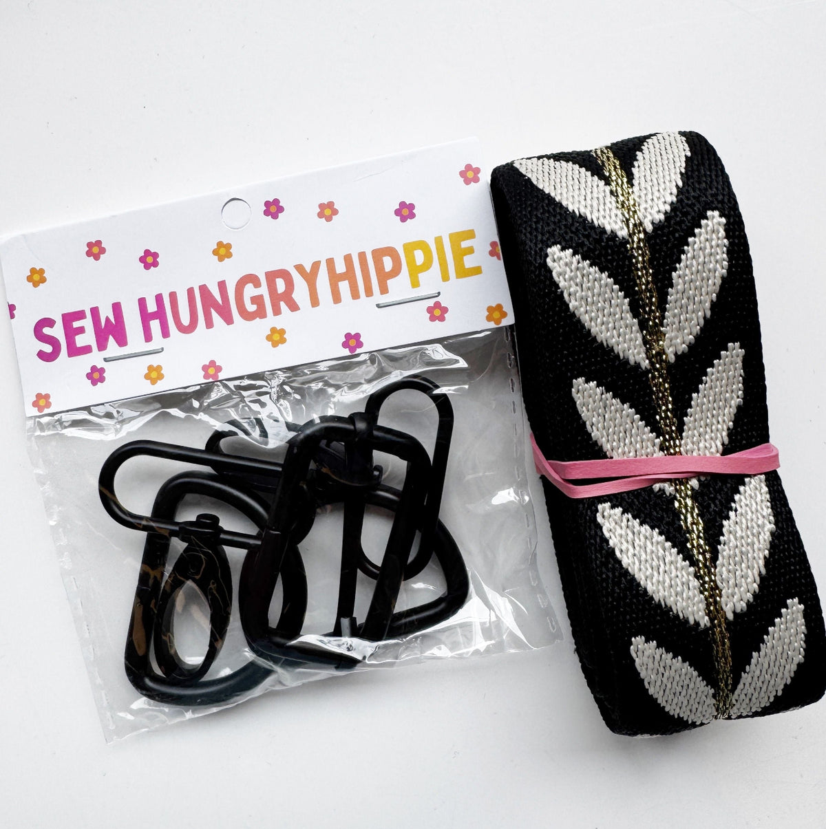 A roll of black and white leaf-patterned jacquard webbing with a metallic gold center line and a package of black hardware clips from Sew Hungry Hippie.