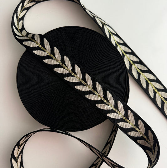 A roll of black webbing with a woven cream and olive green laurel leaf pattern, shown on a clean white background.
