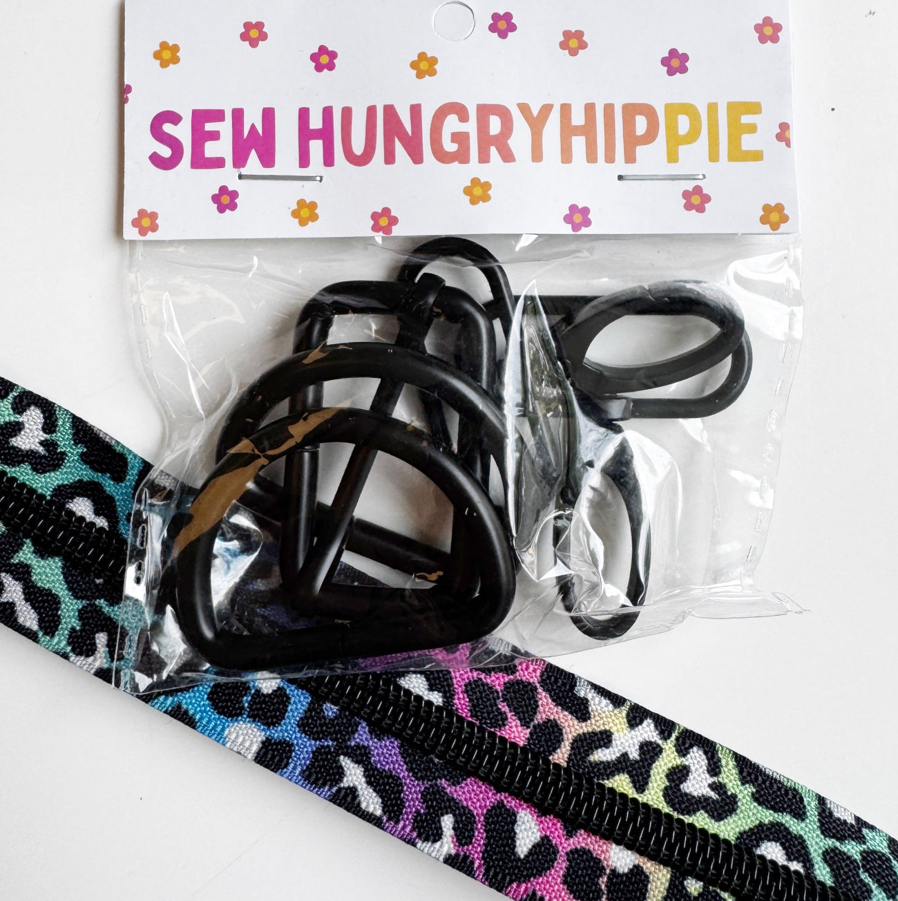 A packaged set of black plastic zipper pulls and a colorful leopard-print zipper tape strip. The items are sealed in a clear plastic bag with a white header card printed with the brand name 'SEW HUNGRY HIPPIE' and small pink and orange flowers.