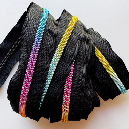 A set of black webbed zippers arranged in a fan shape, each featuring a different metallic zipper track in pink, blue, yellow, and orange, photographed on a plain light background.