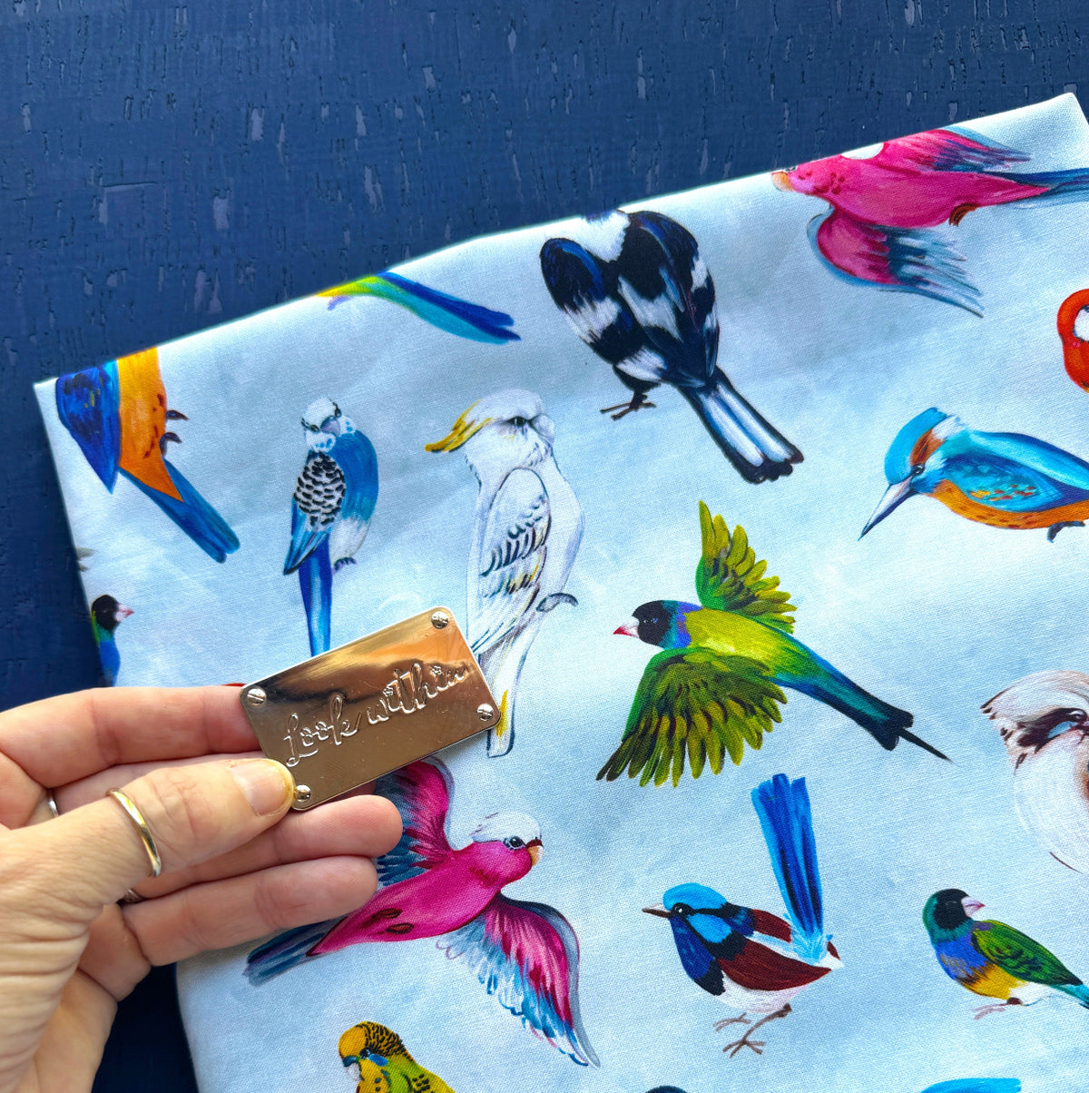 A close-up view of a light blue fabric featuring an illustrated pattern of colorful birds, including a galah, kookaburra, magpie, and various parrots. A hand holding a small, metallic silver rectangle engraved with the cursive text "look within" rests on the lower-left side of the fabric against a dark blue textured background.
