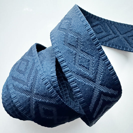 A loose roll of premium slate blue jacquard woven webbing strap featuring an intricate geometric diamond pattern on a white background