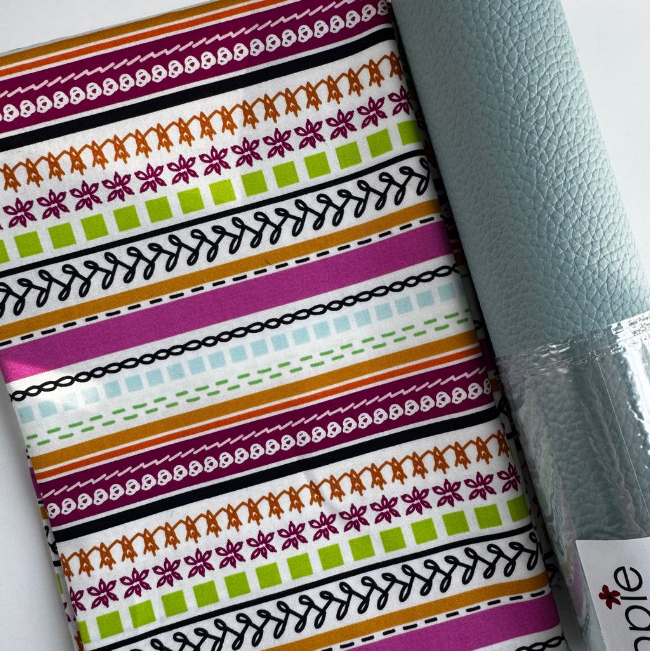 A close-up of folded fabric with horizontal bohemian stripes in purple, orange, and green alongside a roll of pebbled pale blue moonglow vinyl for sewing projects.