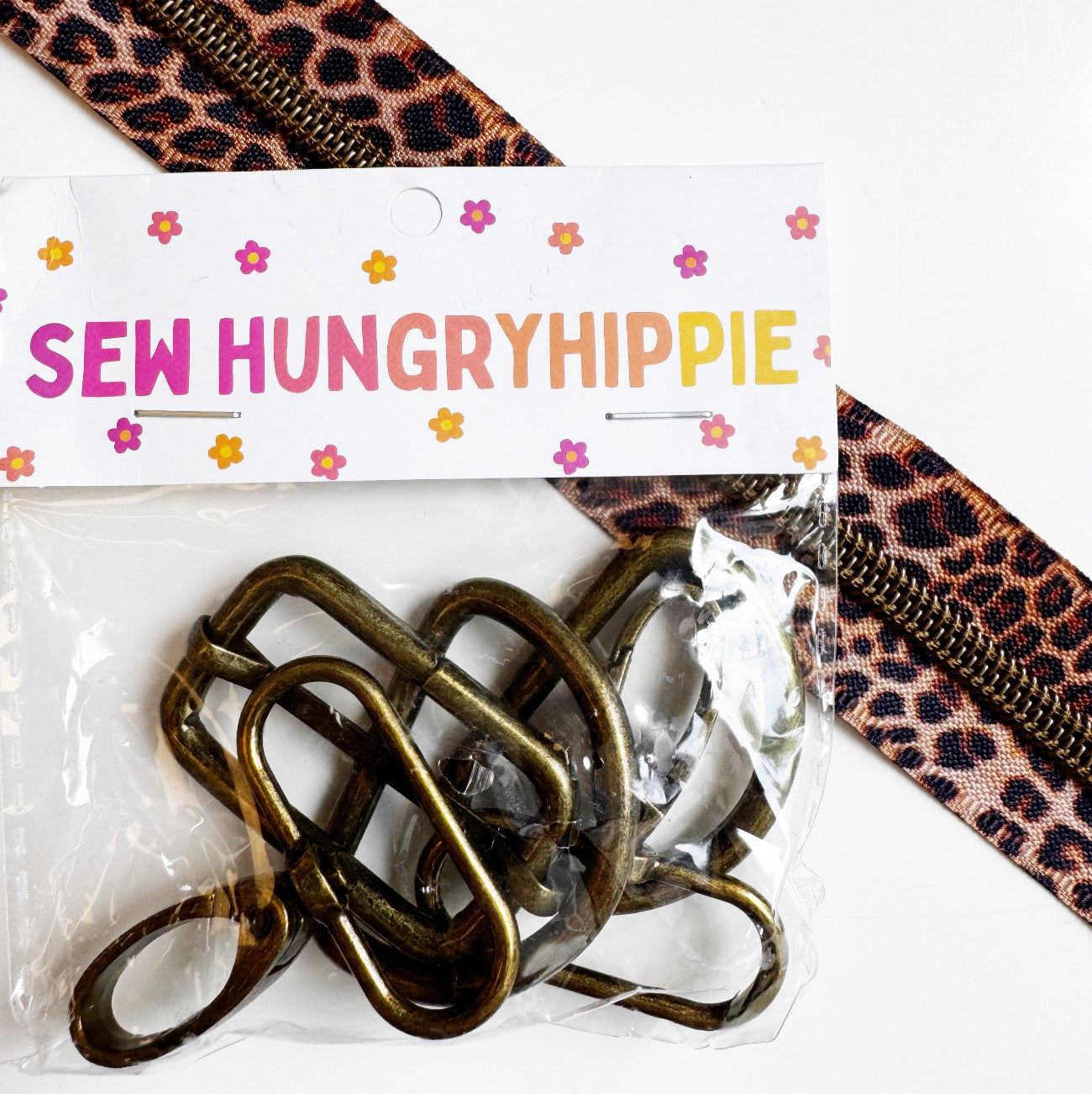A clear plastic bag containing several bronze-toned metal zipper pulls, sealed with a white paper header card printed with the brand name 'SEW HUNGRYHIPPIE' and small colorful flower motifs. A leopard-print zipper tape strip runs diagonally across the top right on a white background.