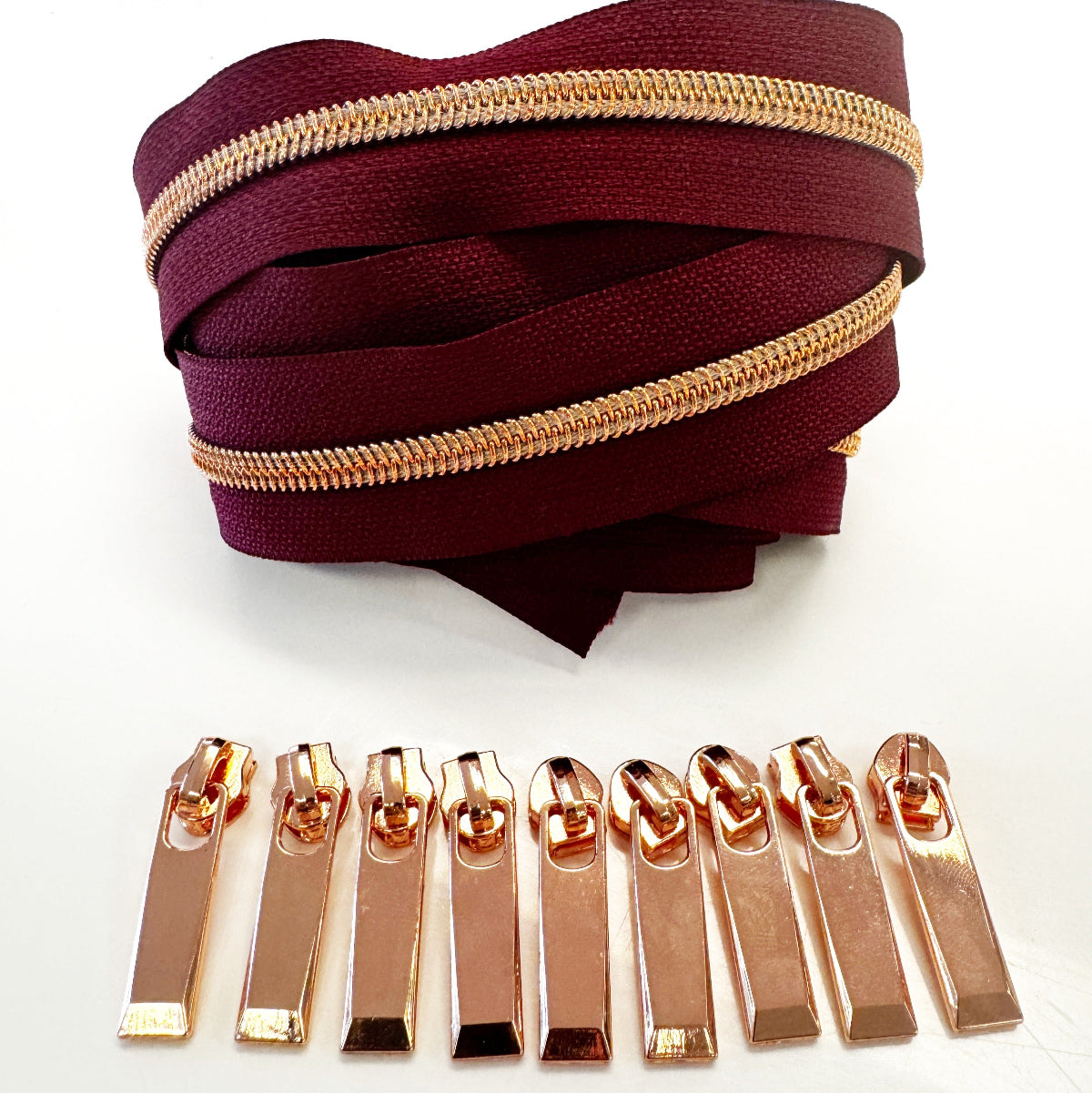 A coiled burgundy zipper ribbon with a metallic rose gold zipper running along the top, displayed above a row of matching rose gold zipper pull tabs on a white background.