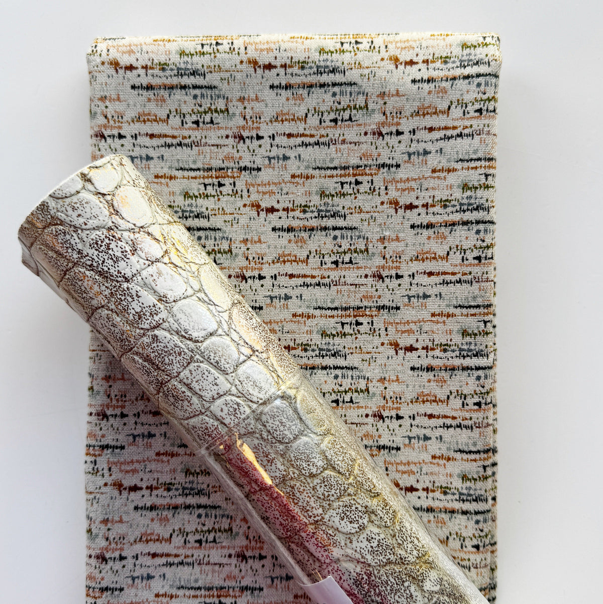 A close-up of a rolled piece of textured fabric with a metallic silver and gold embossed crocodile pattern, laid diagonally over a flat folded sheet of subtle, textured blotted print in a soft eggshell tone patterned material on a white background.