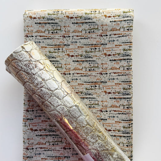 A close-up of a rolled piece of textured fabric with a metallic silver and gold embossed crocodile pattern, laid diagonally over a flat folded sheet of subtle, textured blotted print in a soft eggshell tone patterned material on a white background.