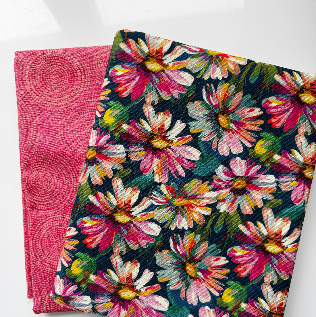 Two folded fabric pieces on a white background: one dark navy floral textile with large pink, white, yellow, and green flowers, and one pink textile with a circular dot pattern.