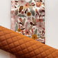 A roll of quilted fabric in a warm, caramel-brown shade layered diagonally across a flat piece of white fabric featuring a vintage-inspired floral pattern with peach, mustard yellow, and brown botanical elements.