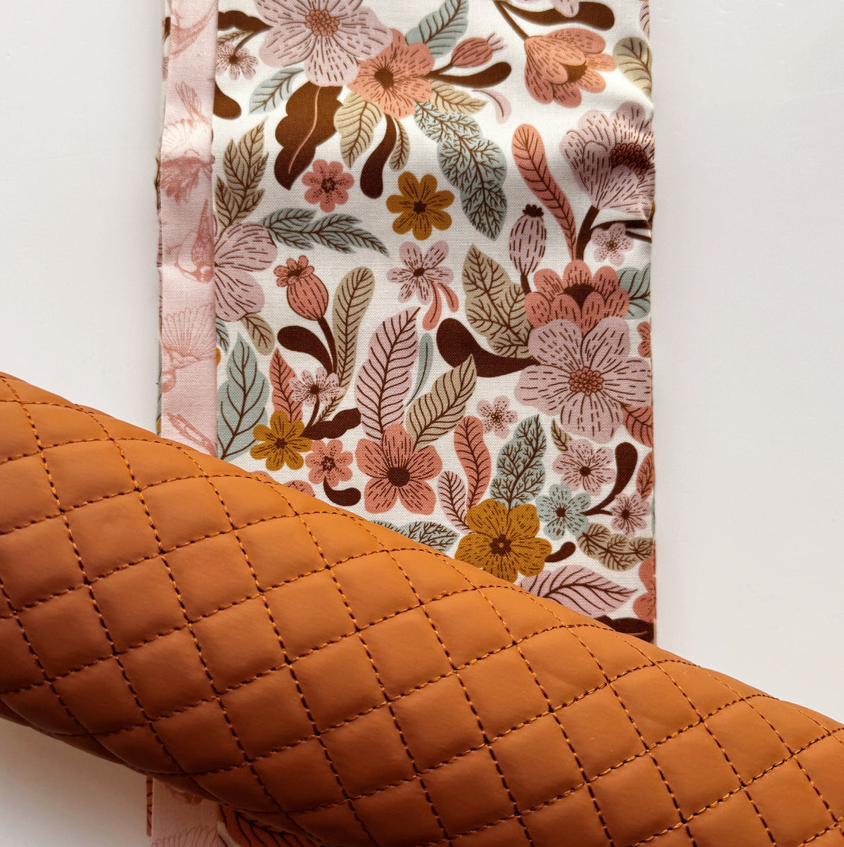 A roll of quilted fabric in a warm, caramel-brown shade layered diagonally across a flat piece of white fabric featuring a vintage-inspired floral pattern with peach, mustard yellow, and brown botanical elements.
