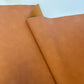 A close-up of a folded piece of smooth, warm tan-brown leather or faux leather showing its texture and color against a light background.
