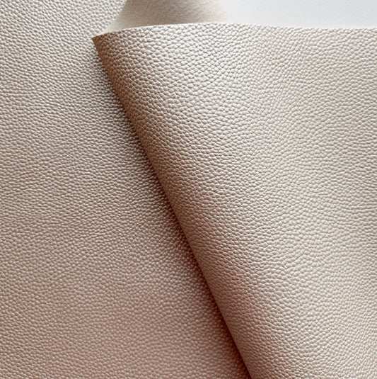 Close-up of champagne-colored Moonglow faux leather vinyl fabric featuring a distinct, finely pebbled texture with a soft metallic sheen, showing the material draped to reveal its smooth backing.