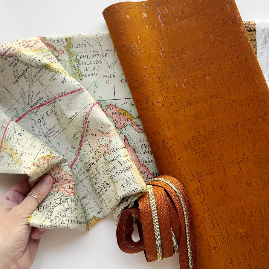 A hand holds a rolled piece of vintage-style map fabric next to a rolled piece of textured tan leather, with a matching tan leather zipper pull and strap shown in front on a white background.