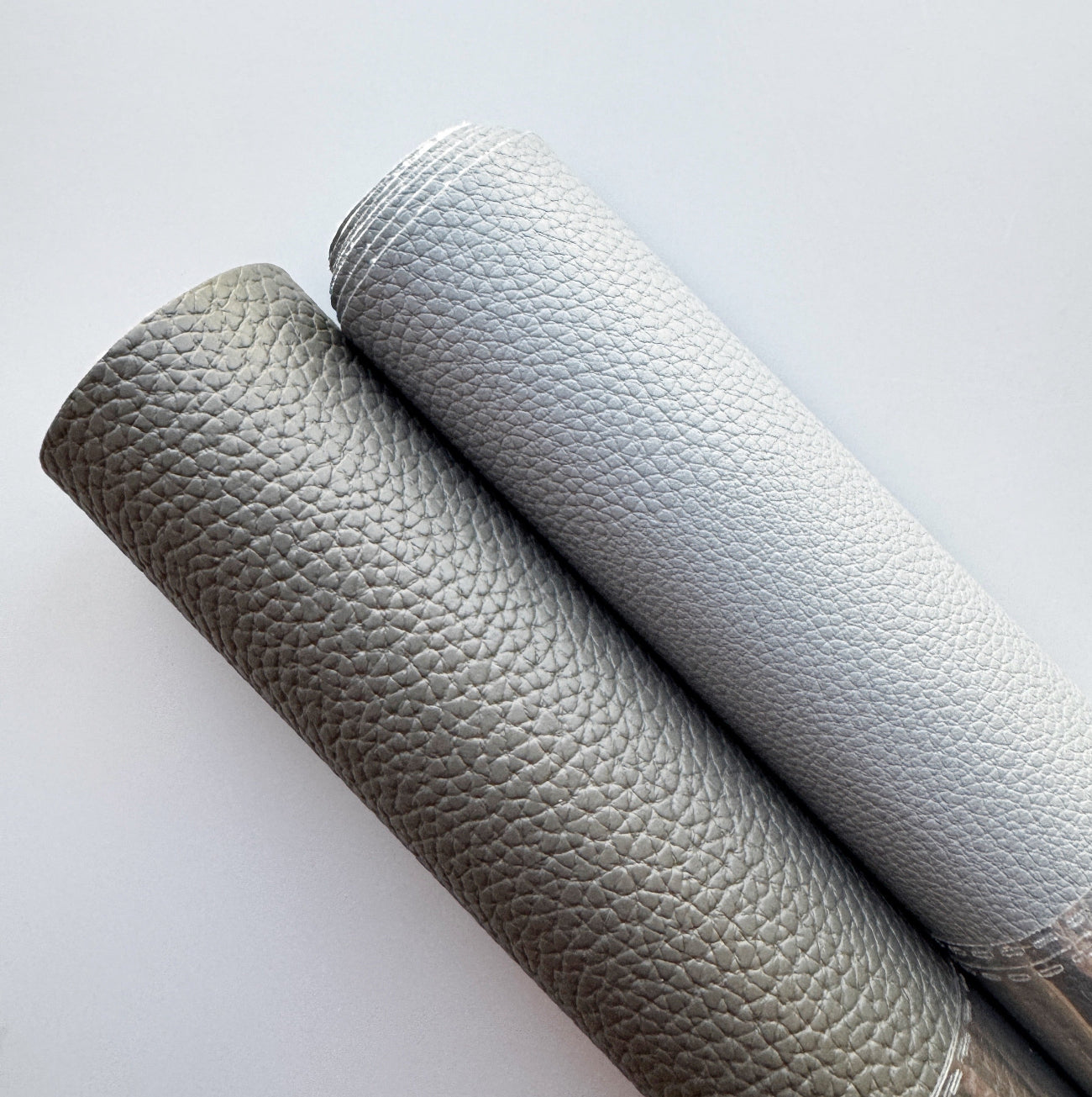 Two rolls of textured faux leather fabric, one in taupe-gray and one in light gray, shown on a clean white background.