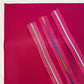 Four strips of bright bright and pink nylon zipper tape with metallic-finish teeth—including silver, gold, and iridescent rainbow colors—are laid diagonally across a sheet of matching textured magenta faux leather fabric.