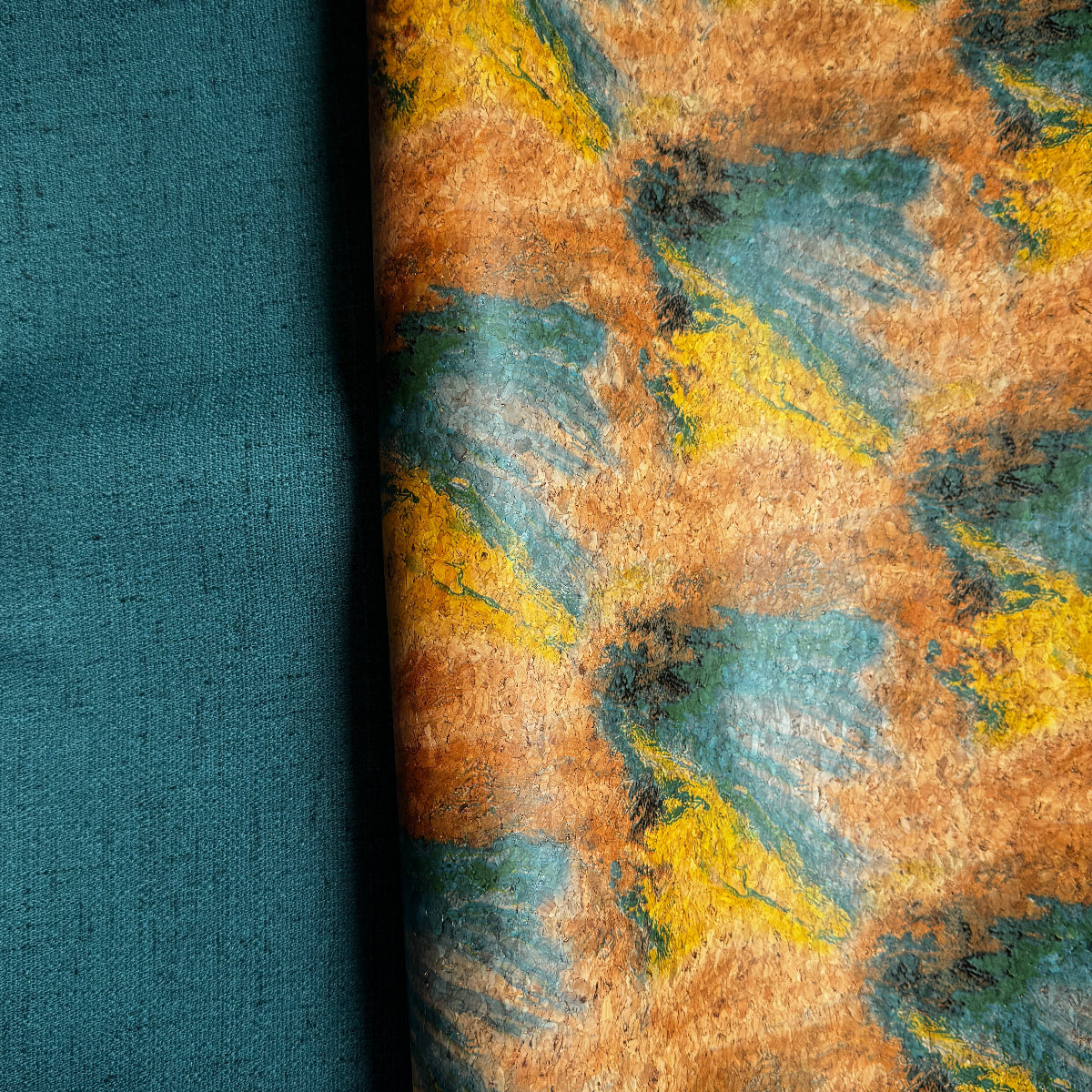 A folded piece of fabric with an abstract brushstroke pattern in mustard yellow, teal, and brown, with a solid teal folded section showing the reverse side, displayed on a white background.