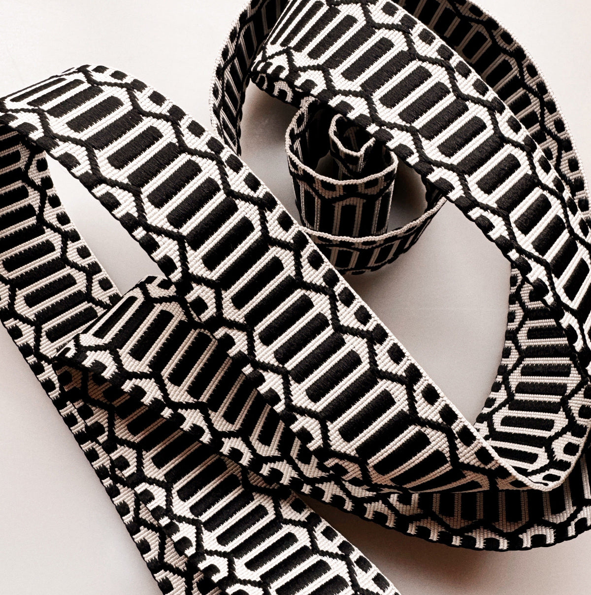 A close-up of a woven ribbon with a bold black and off-white geometric pattern, styled in loops on a light neutral background.