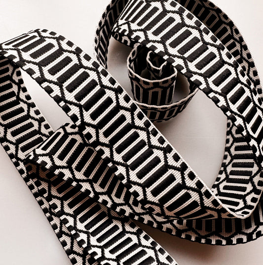 A close-up of a woven ribbon with a bold black and off-white geometric pattern, styled in loops on a light neutral background.