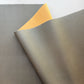 A close-up of a textured faux leather or vinyl material sample in warm gray/taupe, folded over to show a contrasting yellow backing, on a white background.