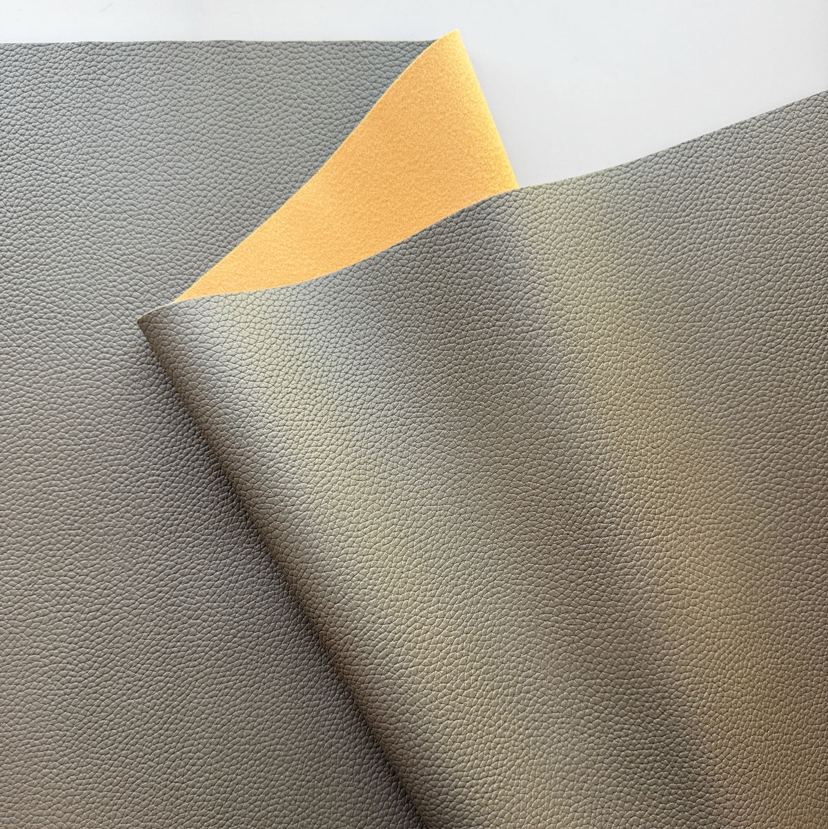 A close-up of a textured faux leather or vinyl material sample in warm gray/taupe, folded over to show a contrasting yellow backing, on a white background.