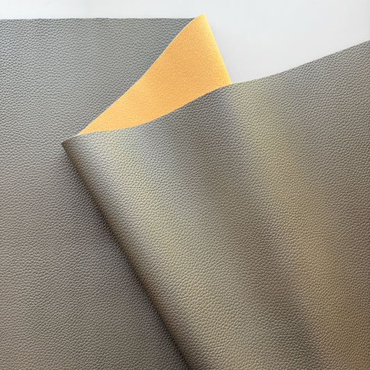 A close-up of a textured faux leather or vinyl material sample in warm gray/taupe, folded over to show a contrasting yellow backing, on a white background.