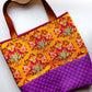 A tote bag with a vibrant floral print on a mustard yellow background, featuring brown leather handles and a quilted purple bottom section.