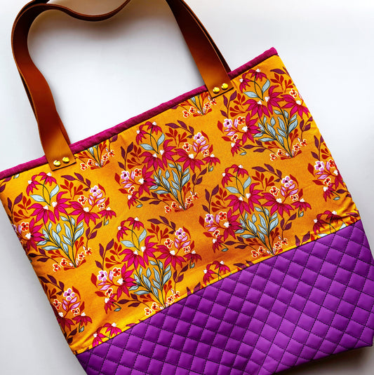 A tote bag with a vibrant floral print on a mustard yellow background, featuring brown leather handles and a quilted purple bottom section.